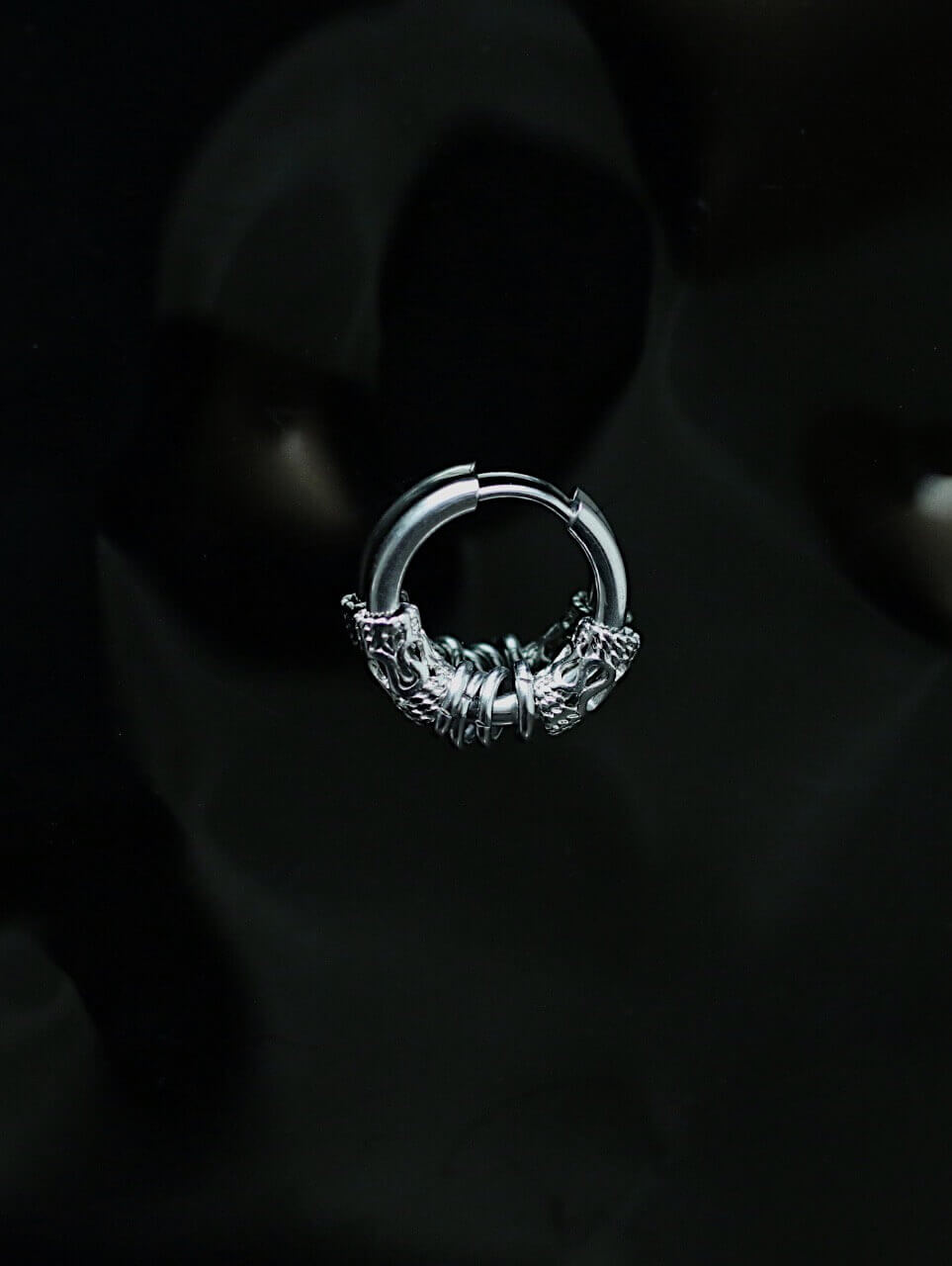 Moody cinematic shot of a tarnish-proof silver gothic hoop earring, aesthetic punk rock jewelry on black reflective surface, hypoallergenic surgical steel.