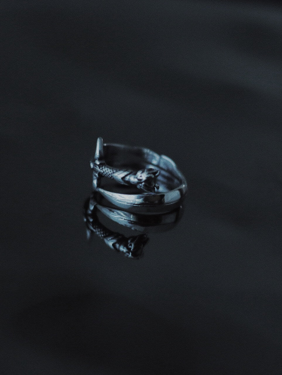 Intricate silver katana sword wrap ring on dark reflective surface, detailed Japanese sword design jewelry, tarnish-proof stainless steel amulet, Monora Studio.