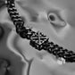 Close up shot of Monora Studio industrial gothic geometric cross monogram necklace on a fluid metallic background