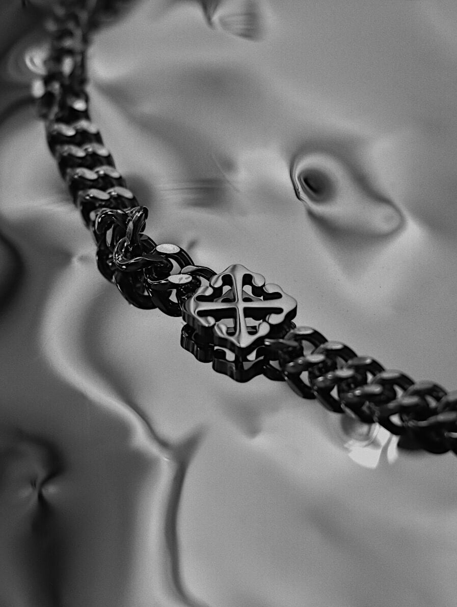 Close up shot of Monora Studio industrial gothic geometric cross monogram necklace on a fluid metallic background