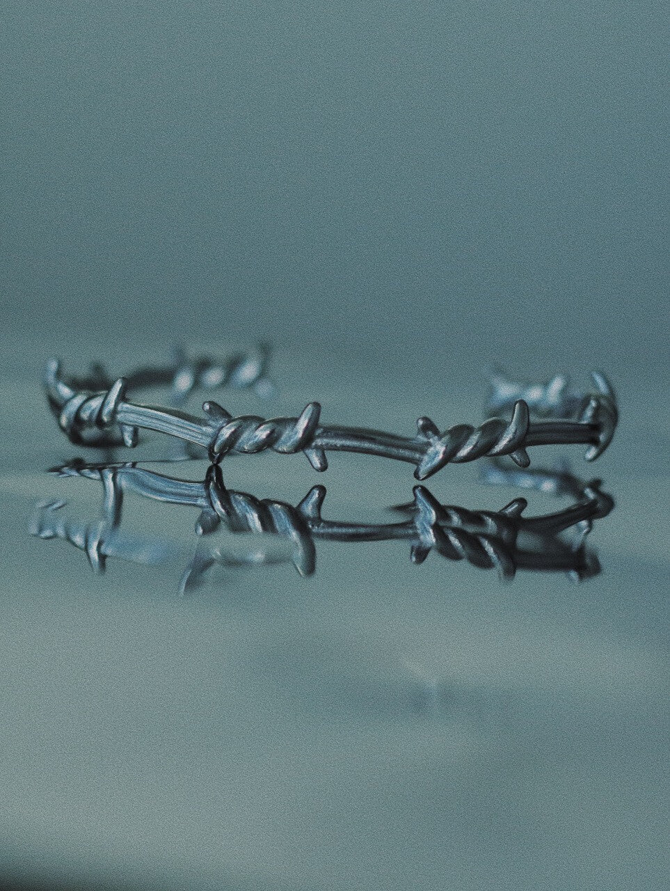 Monora Studio stainless steel barbed wire cuff bracelet reflecting on dark glass water surface close up⁠
