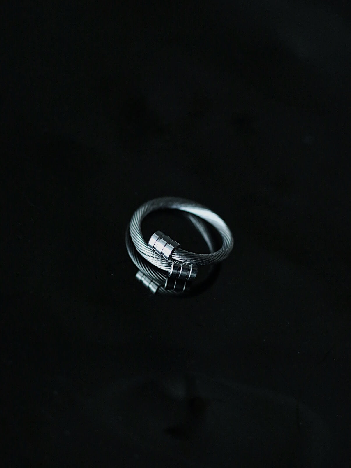 High-end product shot of the stainless steel cable bypass ring on a moody black background with realistic reflection, premium tarnish-proof silver wire ring, unisex streetwear accessory.